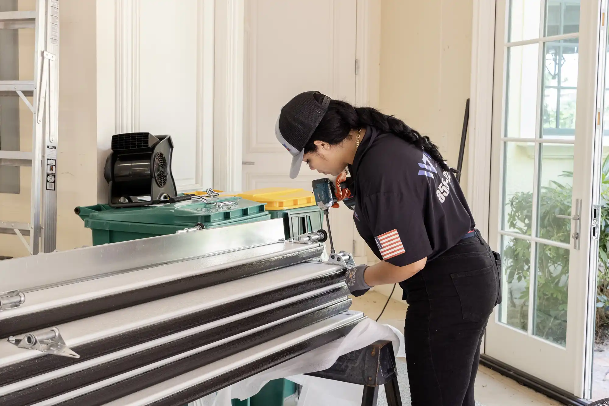 Female technician inspecting and assembling garage door panels using a power drill on a workbench in a residential garage.