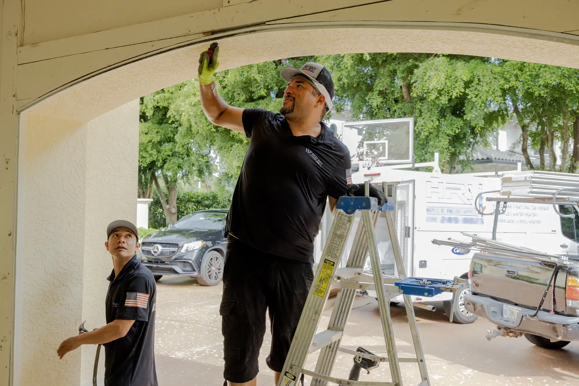 Technician standing on a ladder fixing the curved track of a garage door while another technician observes, with vehicles parked outside.