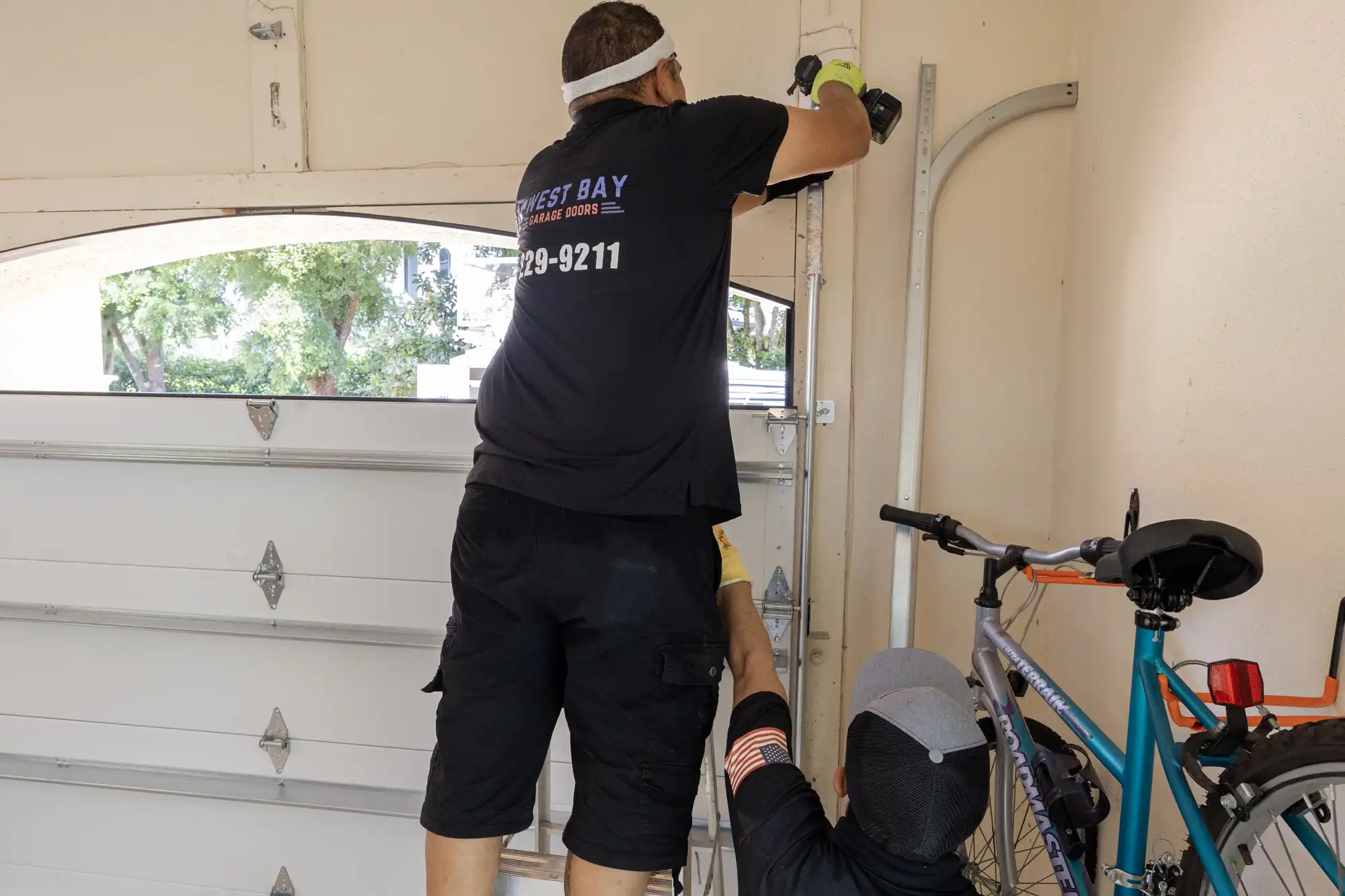 Two technicians installing a garage door, with one using a power drill and the other assisting, inside a residential garage with a bicycle nearby