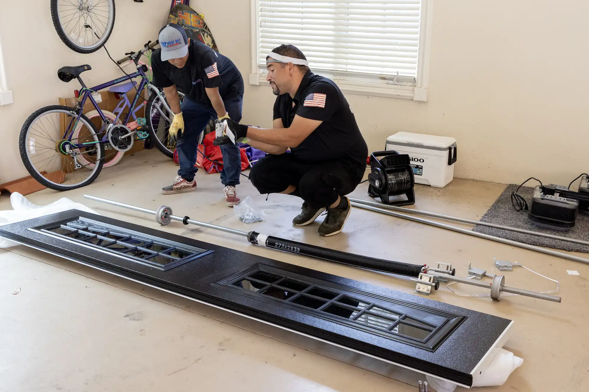 Two technicians in a garage preparing to install a garage door, with tools and components laid out on the floor.