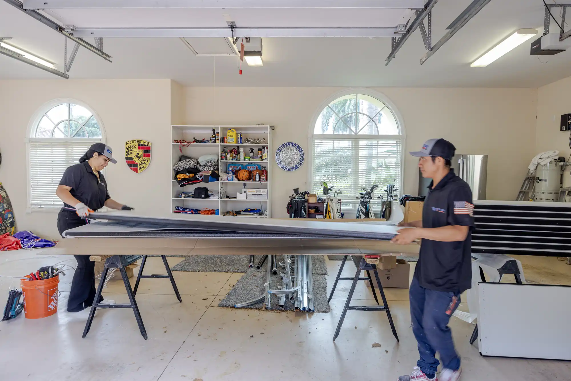 Two technicians in a garage working together to assemble a garage door panel on sawhorses, surrounded by tools and equipment.